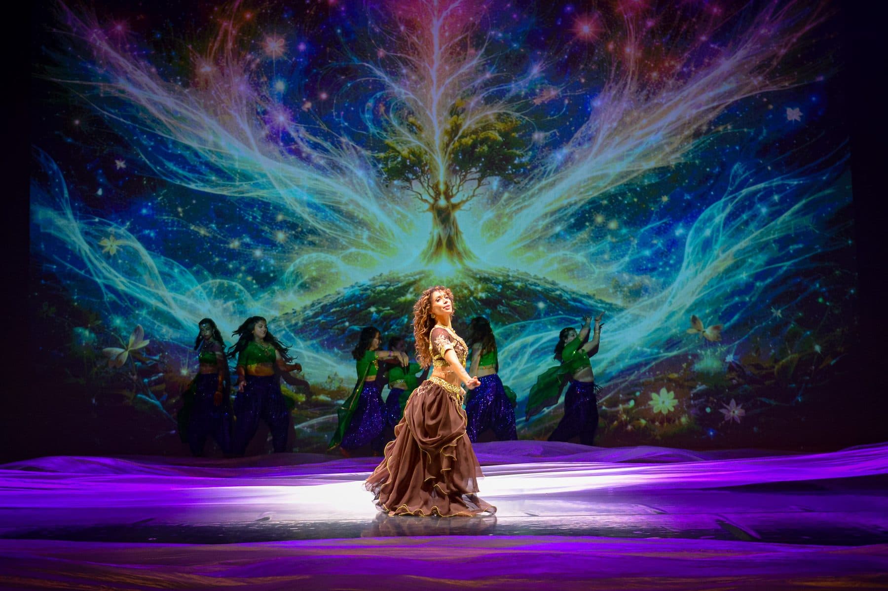 Tableau de spectacle devant un arbre de lumière projeté, danseuses en costumes clairs